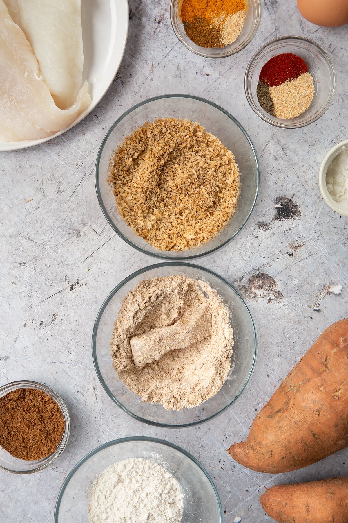 Two bowls, one containing breadcrumbs mixed with spices and another containing flour mixed with spices. A piece of cod is dipped in the flour. Surrounding the bowls is ingredients for spicy fish sticks.