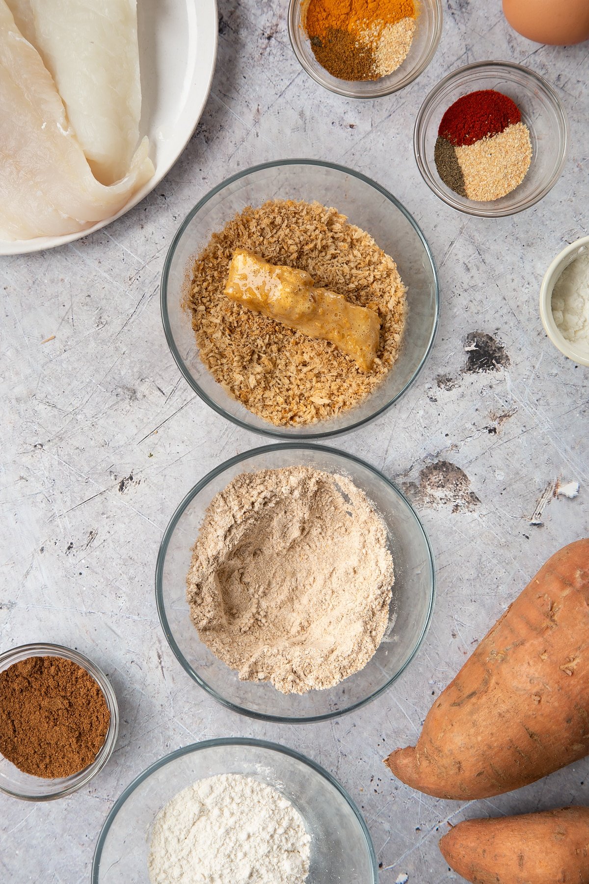 Two bowls, one containing breadcrumbs mixed with spices and another containing flour mixed with spices. A piece of cod is dipped in the breadcrumbs. Surrounding the bowls is ingredients for spicy fish sticks.