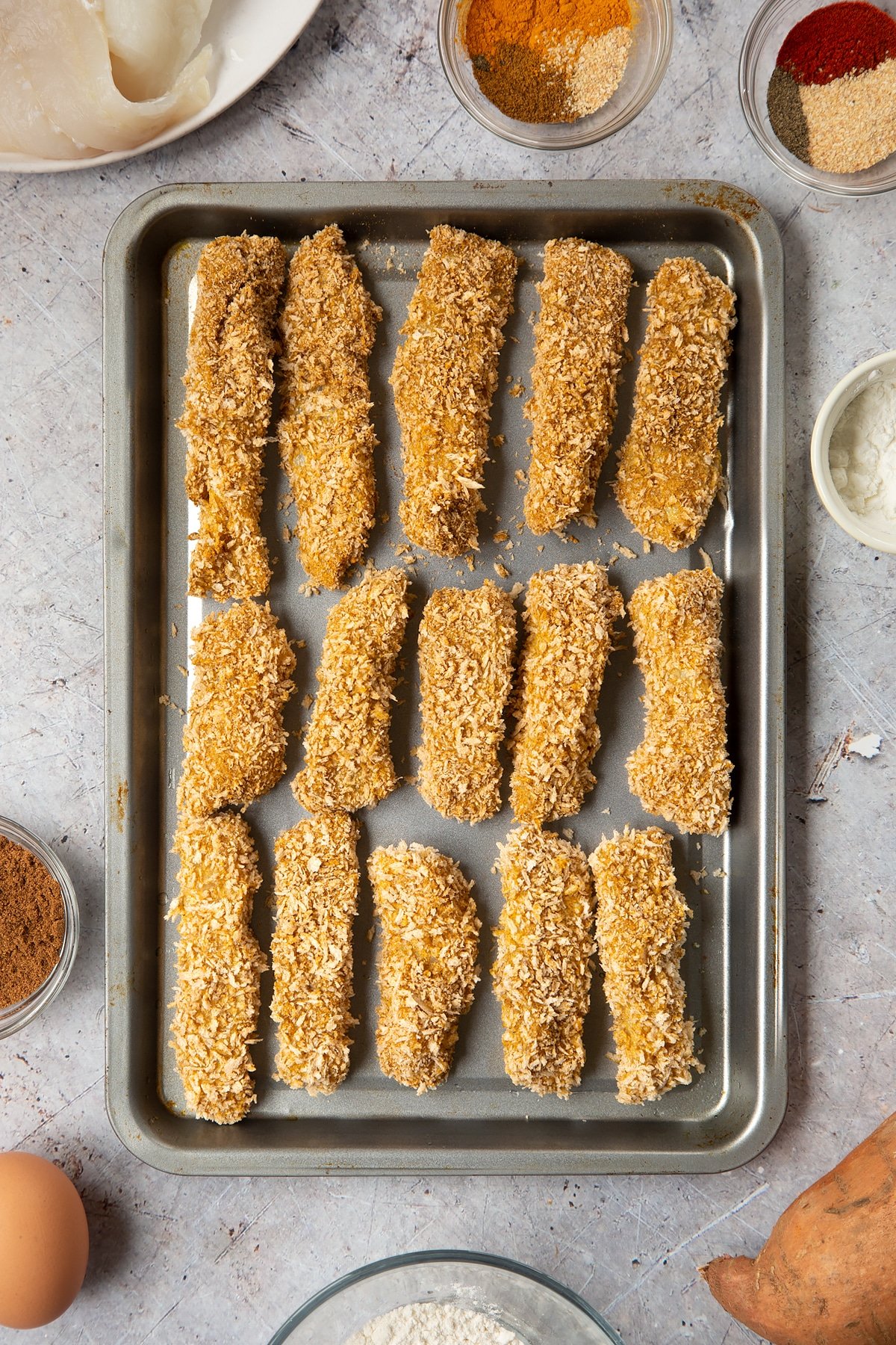 Breaded fish sticks arranged on a baking tray. Surrounding the tray is ingredients for spicy fish sticks.