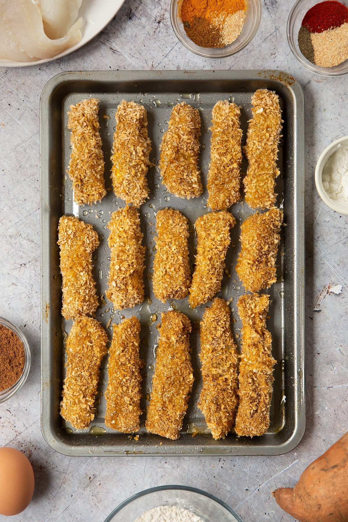 Breaded fish sticks arranged on a baking tray, sprayed with oil. Surrounding the tray is ingredients for spicy fish sticks.