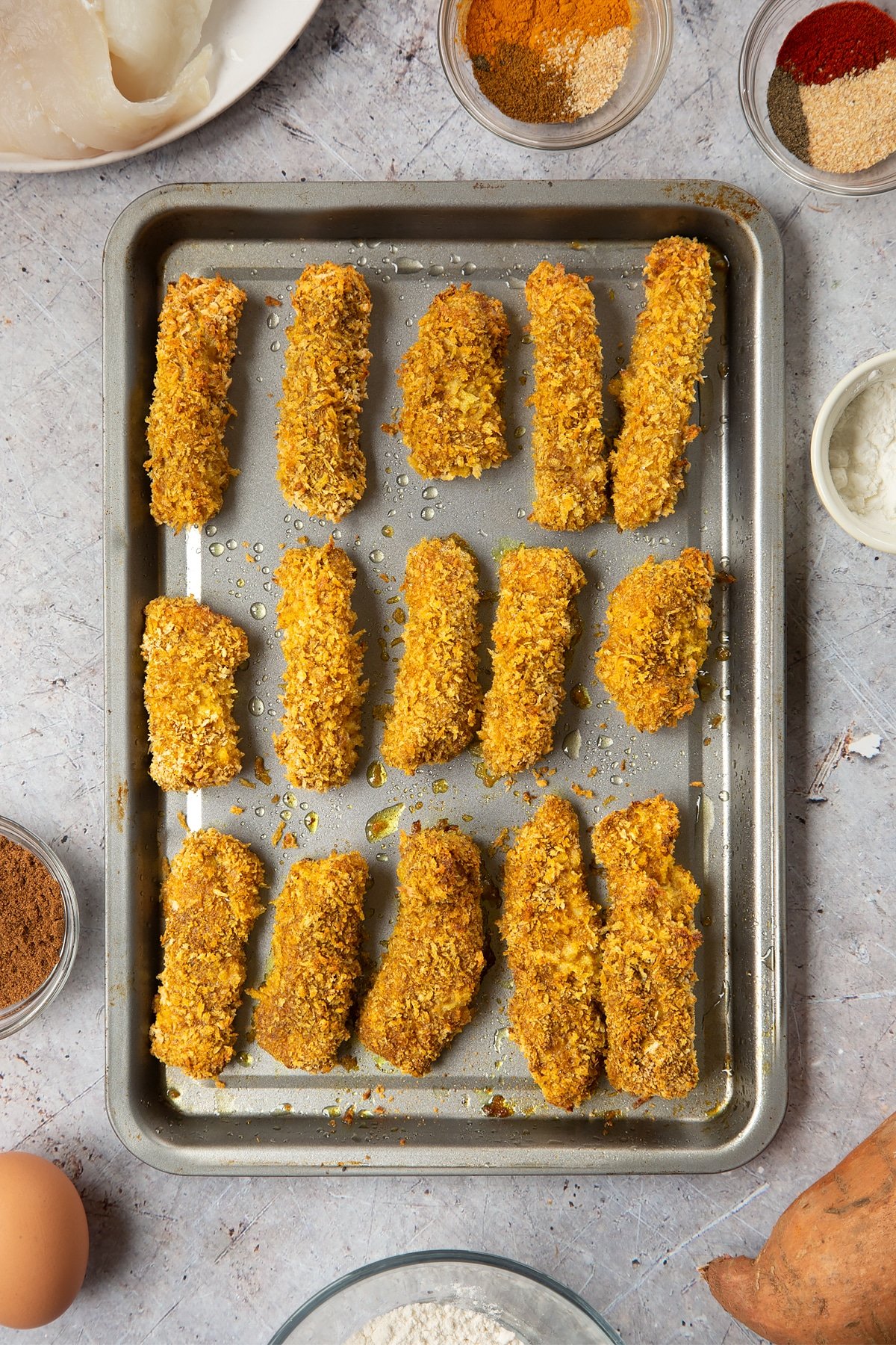 Breaded fish sticks arranged on a baking tray and freshly baked and golden. Surrounding the tray is ingredients for spicy fish sticks.