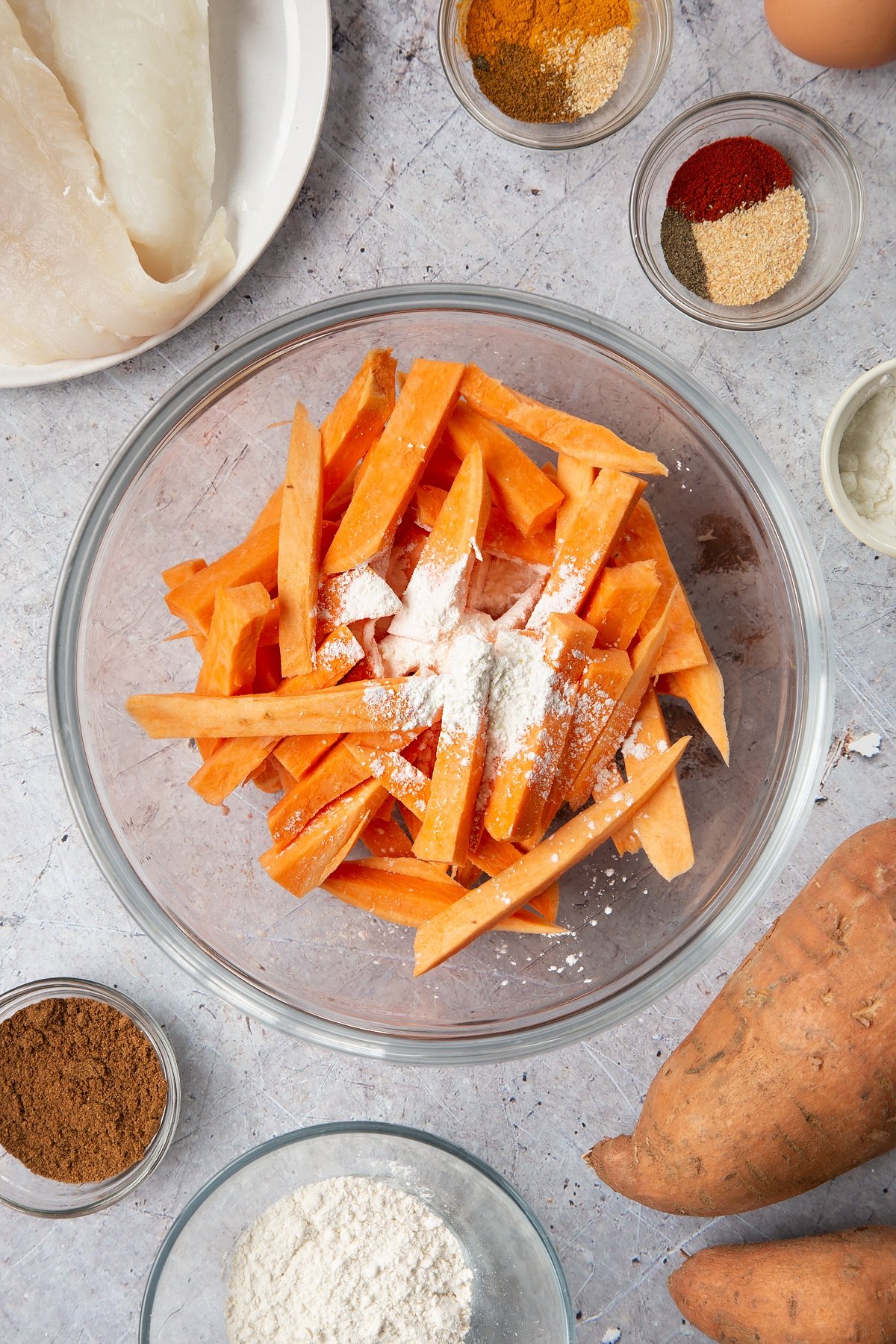Sweet potato cut into fries and placed in a glass bowl, topped with cornflour. Surrounding the bowl is ingredients for sweet potato chips.