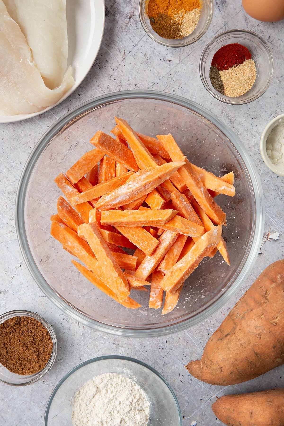 Sweet potato cut into fries and tossed in cornflour in a glass bowl. Surrounding the bowl is ingredients for sweet potato chips.