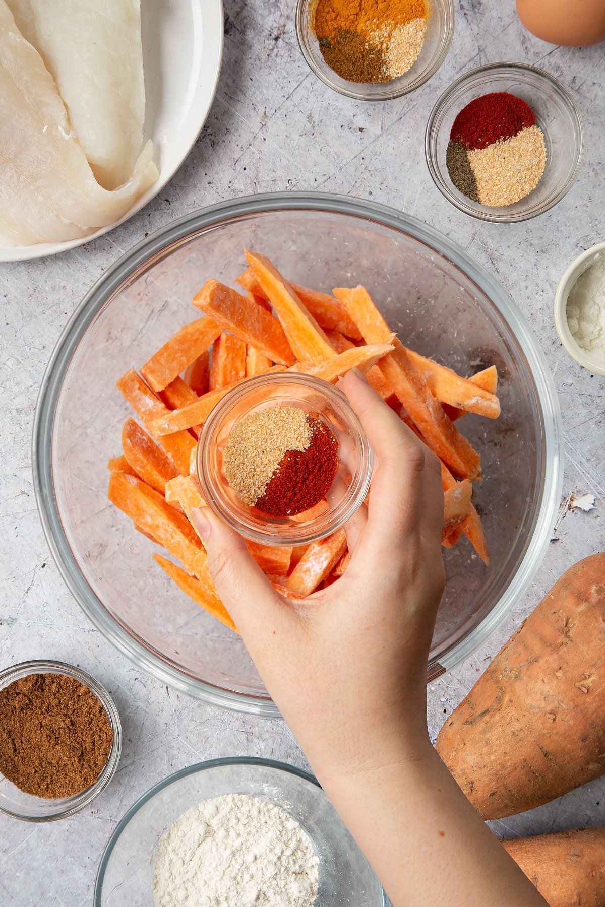 Sweet potato cut into fries and tossed in cornflour in a glass bowl. A hand holds a small bowl containing garlic granules and paprika. Surrounding the bowl is ingredients for sweet potato chips.