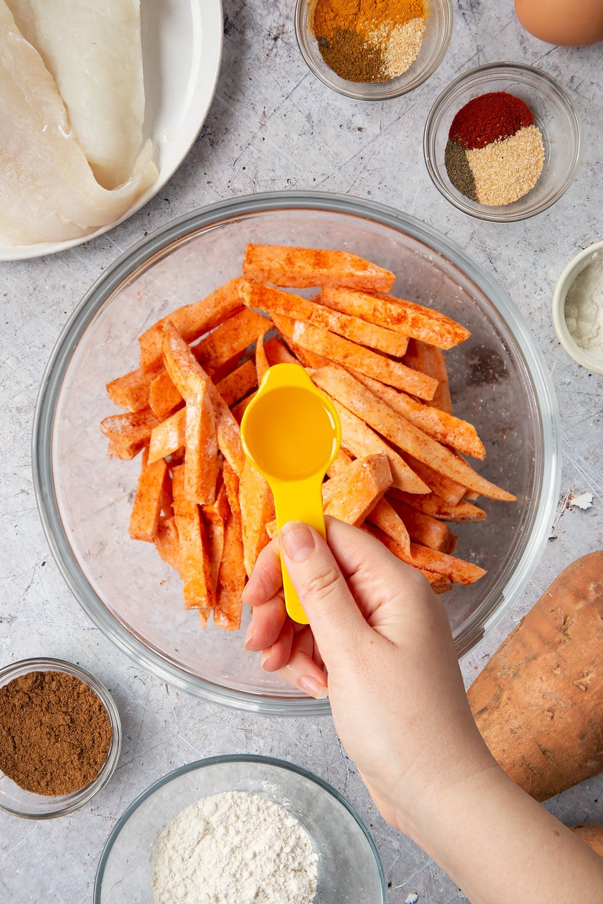 Sweet potato cut into fries and tossed in cornflour in a glass bowl. A hand holds a tablespoon of oil above the bowl. Surrounding the bowl is ingredients for sweet potato chips.