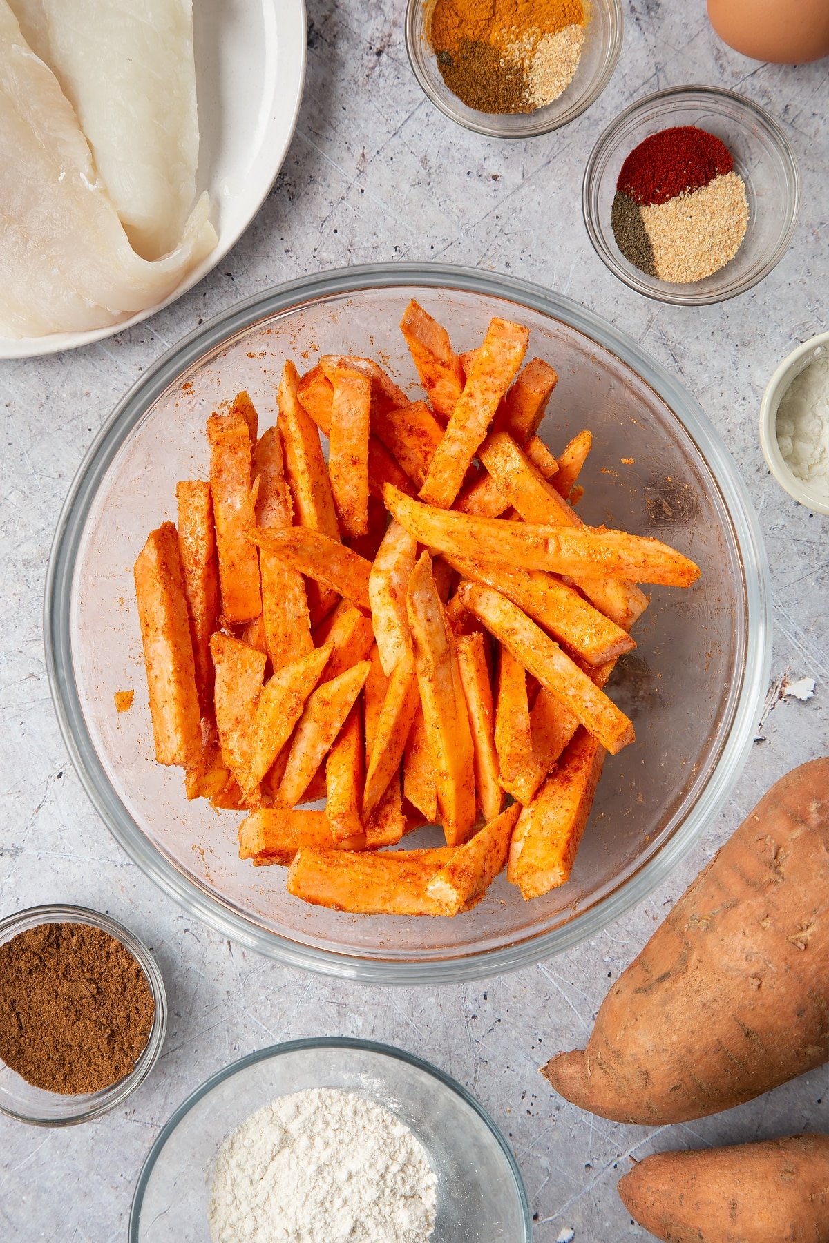 Sweet potato in a bowl. It has been cut into fries and tossed in cornflour, garlic granules, paprika and oil. Surrounding the bowl is ingredients for sweet potato chips.