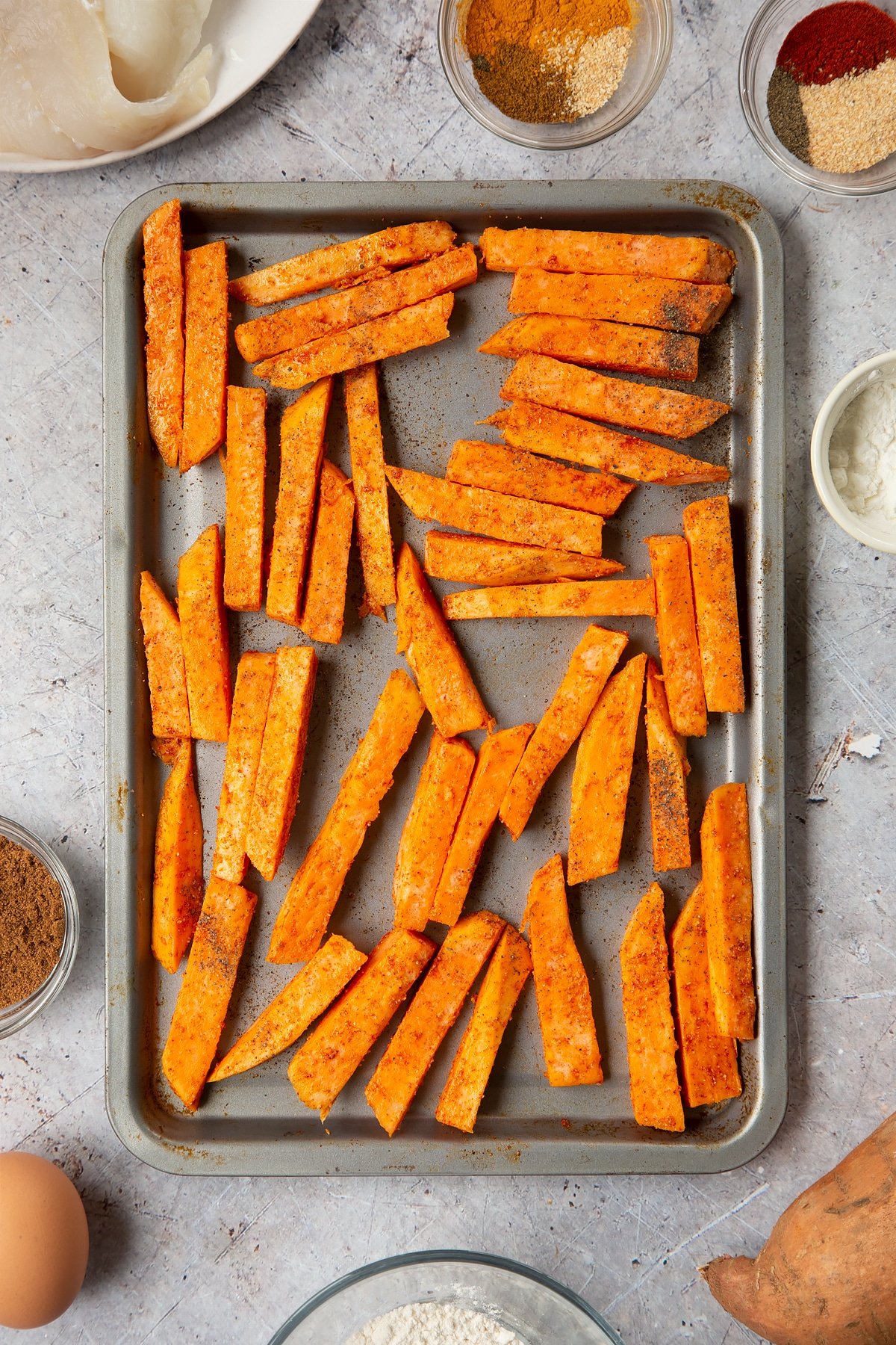 Sweet potato chips, tossed in cornflour, garlic granules, paprika and oil, arranged on a tray ready to be cooked. Surrounding the tray is ingredients for sweet potato chips.