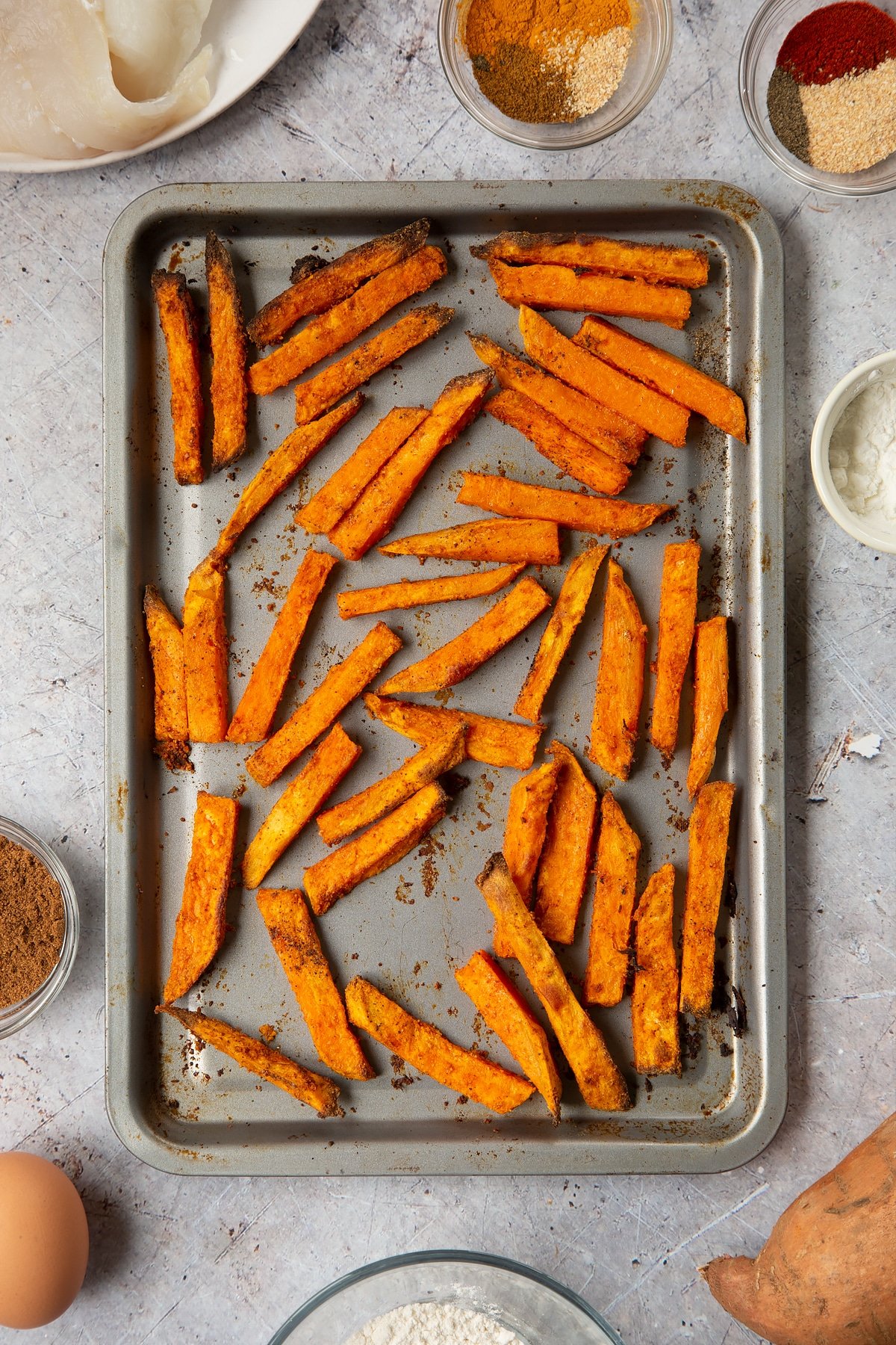 Sweet potato chips, tossed in cornflour, garlic granules, paprika and oil, arranged on a tray and just baked. Surrounding the tray is ingredients for sweet potato chips.