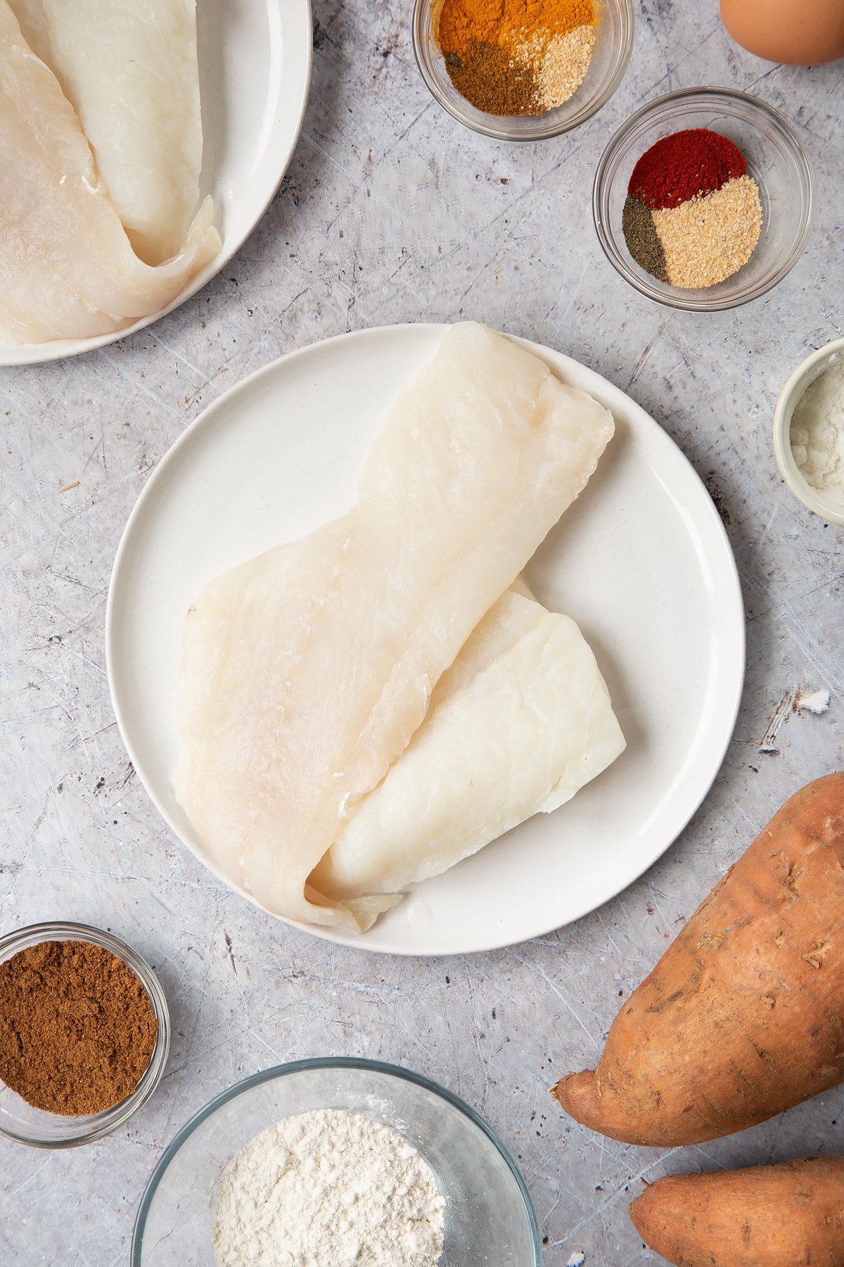 Two cod fillets on a white plate. Surrounding the plate is ingredients for spicy fish sticks.