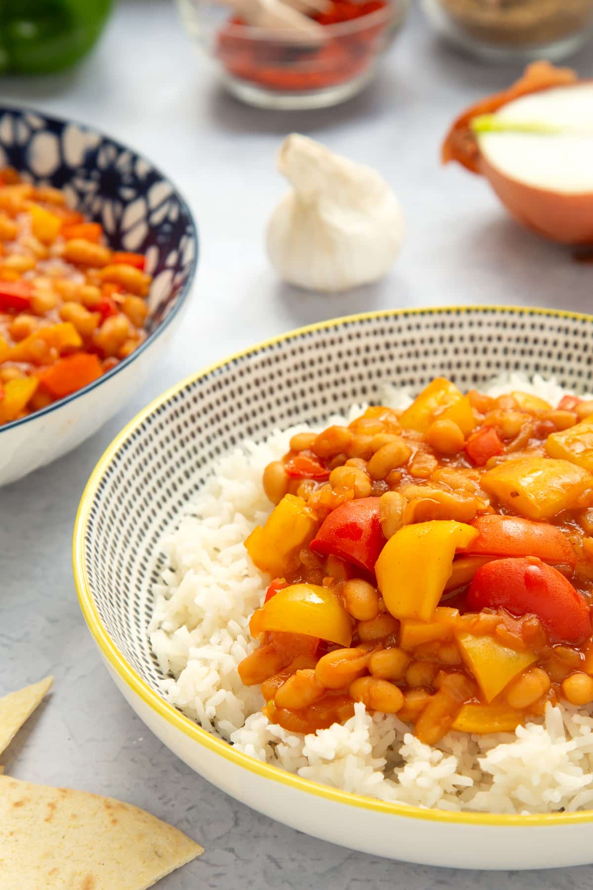 A 5 minute baked bean chilli served on a bed of rice in a bowl. More chilli is in a bowl to the background and ingredients to make the chilli surround the bowls.