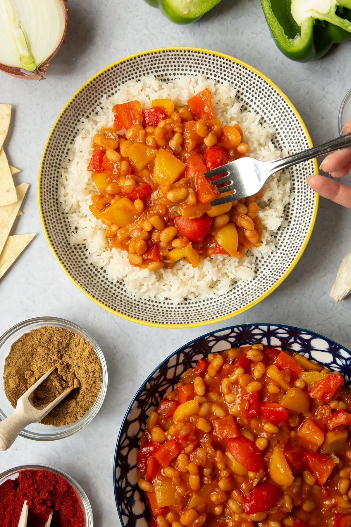 A 5 minute baked bean chilli served on a bed of rice in a bowl. A fork lifts a bite of chilli. More chilli is in a bowl to the background and ingredients to make the chilli surround the bowls.