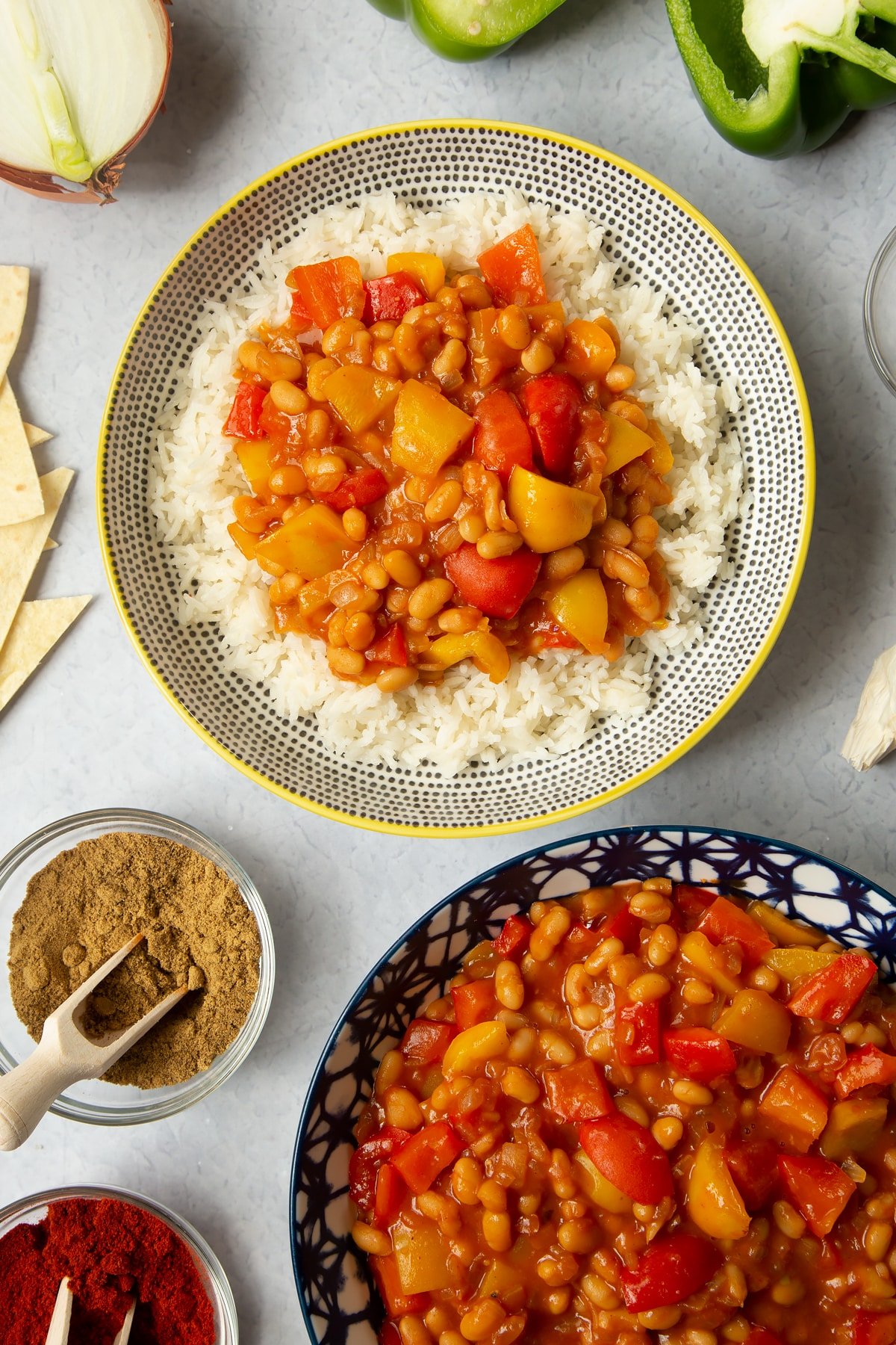 A 5 minute baked bean chilli served on a bed of rice in a bowl. More chilli is in a bowl to the side and ingredients to make the chilli surround the bowls.