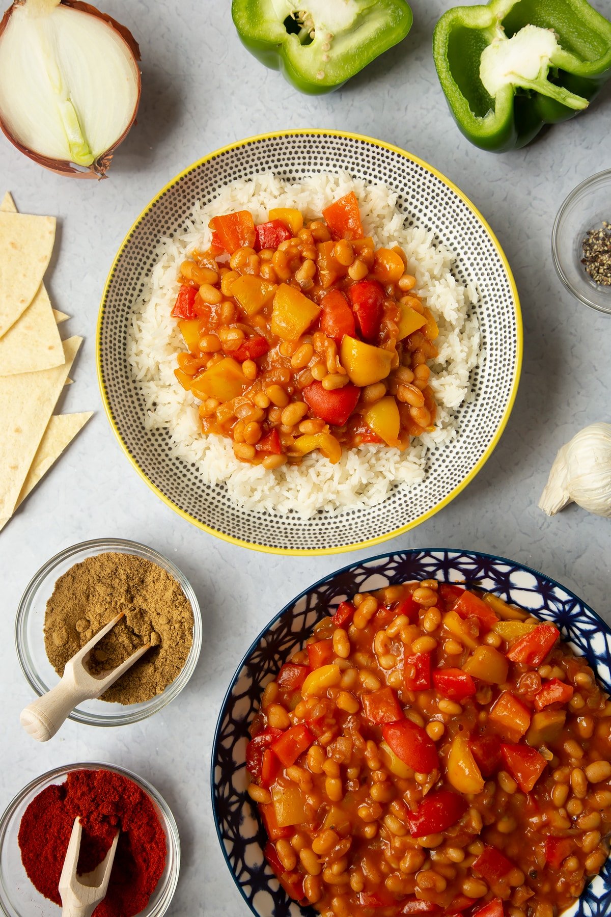 Baked bean chilli served on rice. Another bowl holds more chilli. Ingredients such as peppers, garlic and spices surround the bowls.