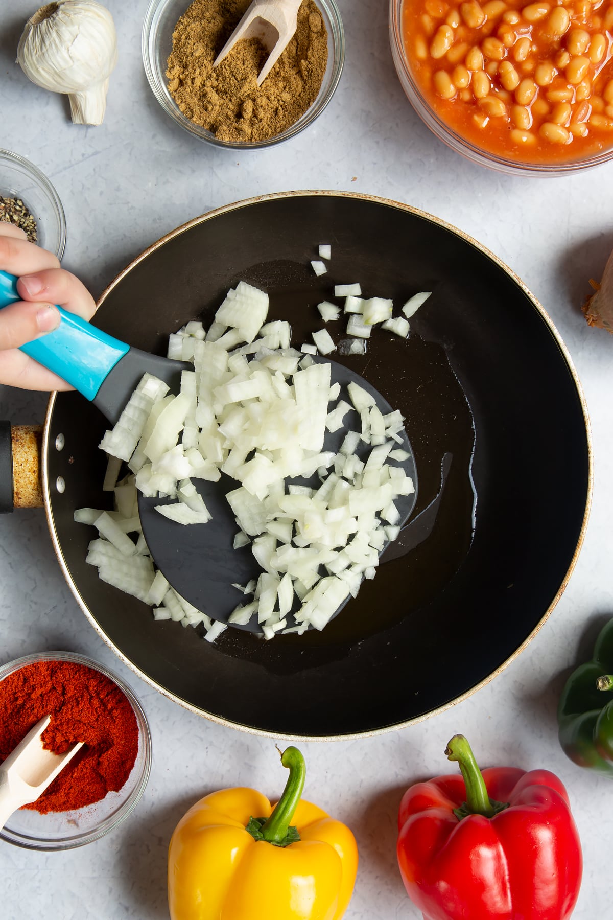 A frying pan containing oil. Above the pan, a hand is adding chopped onion. Ingredients to make baked bean chilli surround the pan.