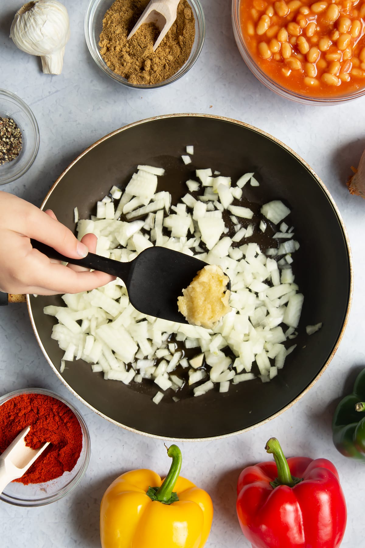 A frying pan containing oil and onion. Above the pan, a hand is adding chopped garlic. Ingredients to make baked bean chilli surround the pan.