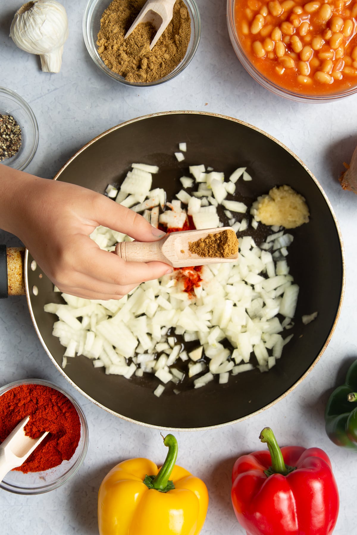 A frying pan containing oil, onion, garlic and paprika. Above the pan, a hand is adding cumin. Ingredients to make baked bean chilli surround the pan.