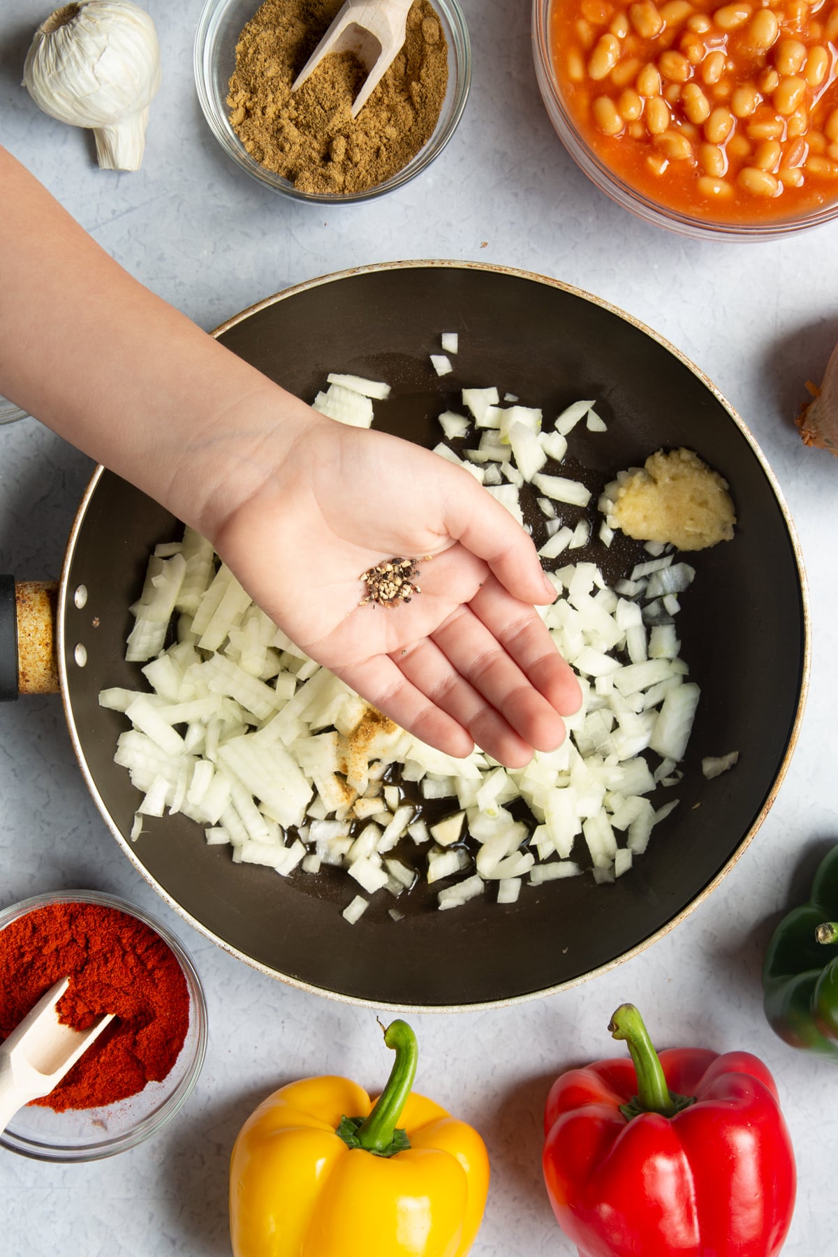 A frying pan containing oil, onion, garlic, paprika and cumin. Above the pan, a hand is adding black pepper. Ingredients to make baked bean chilli surround the pan.