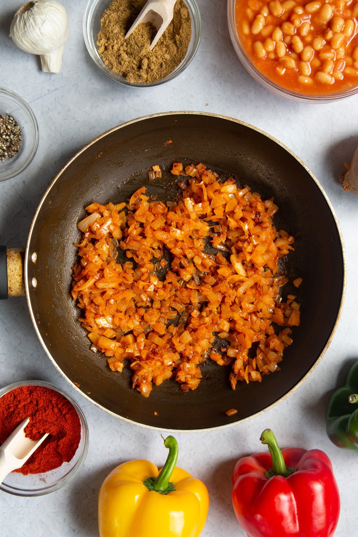 A frying pan containing fried onion, garlic, paprika, cumin and pepper. Ingredients to make baked bean chilli surround the pan.