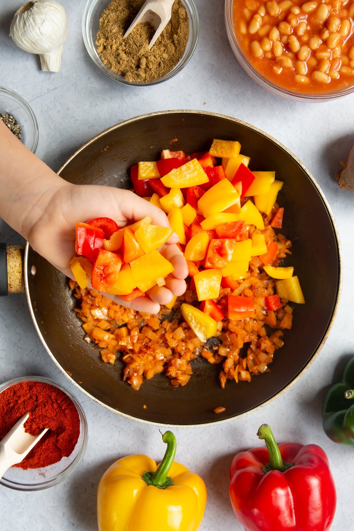 A frying pan containing fried onion, garlic, paprika, cumin and pepper. A hand is adding fresh chopped peppers. Ingredients to make baked bean chilli surround the pan.