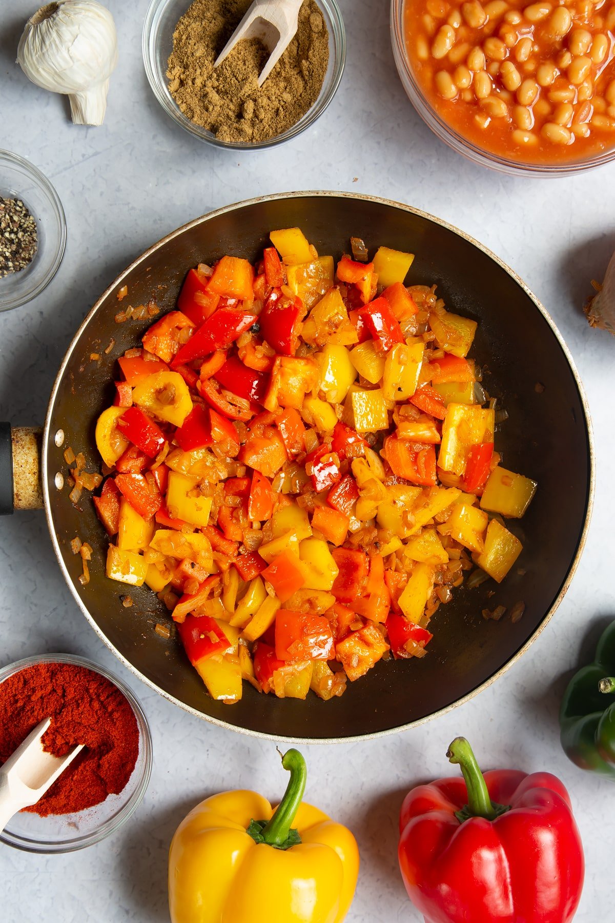 A frying pan containing fried onion, garlic, paprika, cumin, pepper and red and yellow peppers. Ingredients to make baked bean chilli surround the pan.