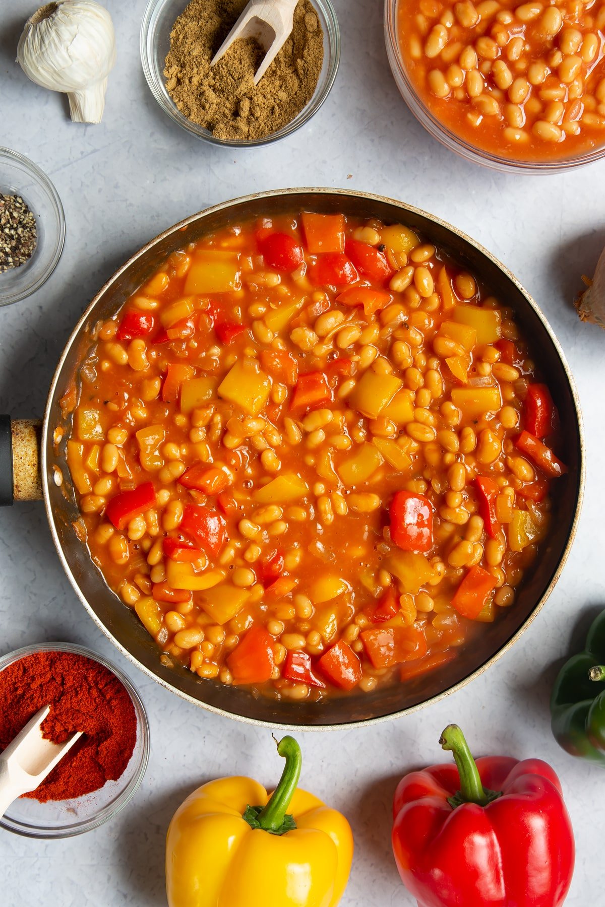 A frying pan containing fried onion, garlic, paprika, cumin, pepper and red and yellow peppers and baked beans. Ingredients to make baked bean chilli surround the pan.