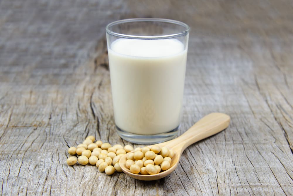 A glass of soy milk on a rustic wooden table. In front of the glass is some soy beans on a wooden spoon. 