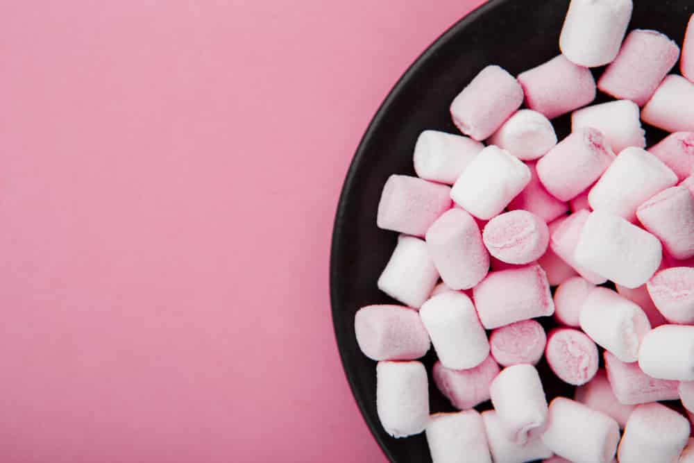 Small pink and white marshmallows in  a black bowl on a pink background. 