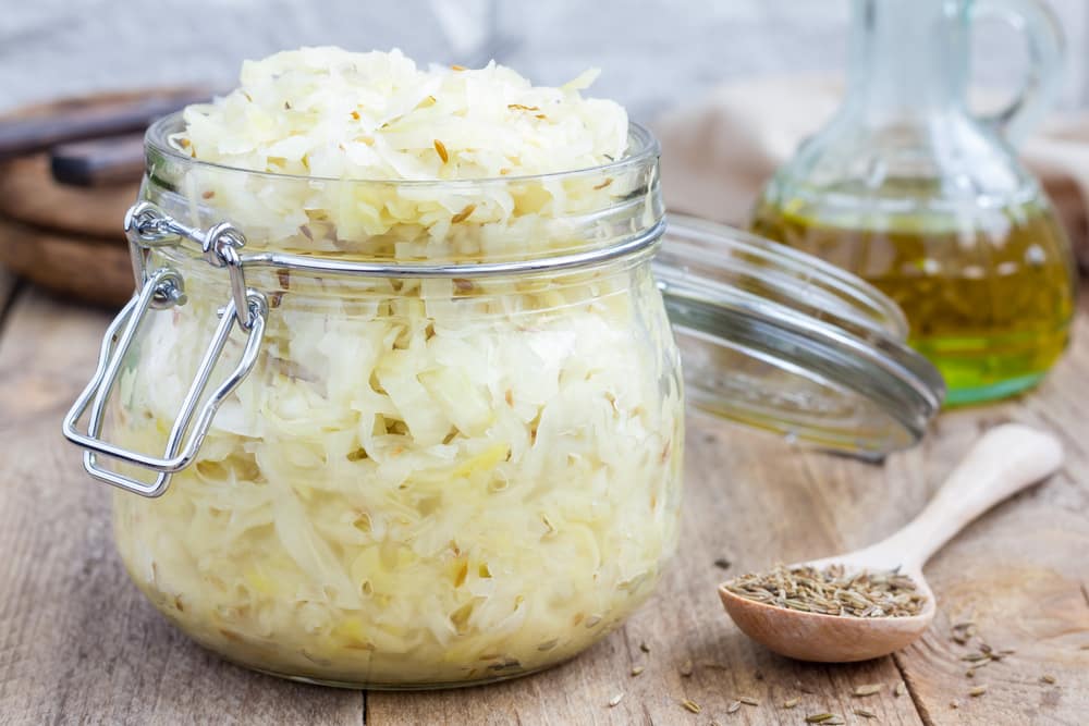 A jar filled with sauerkraut. The lid is open. At the side of the jar there's a couple of ingredients sat on a wooden table.