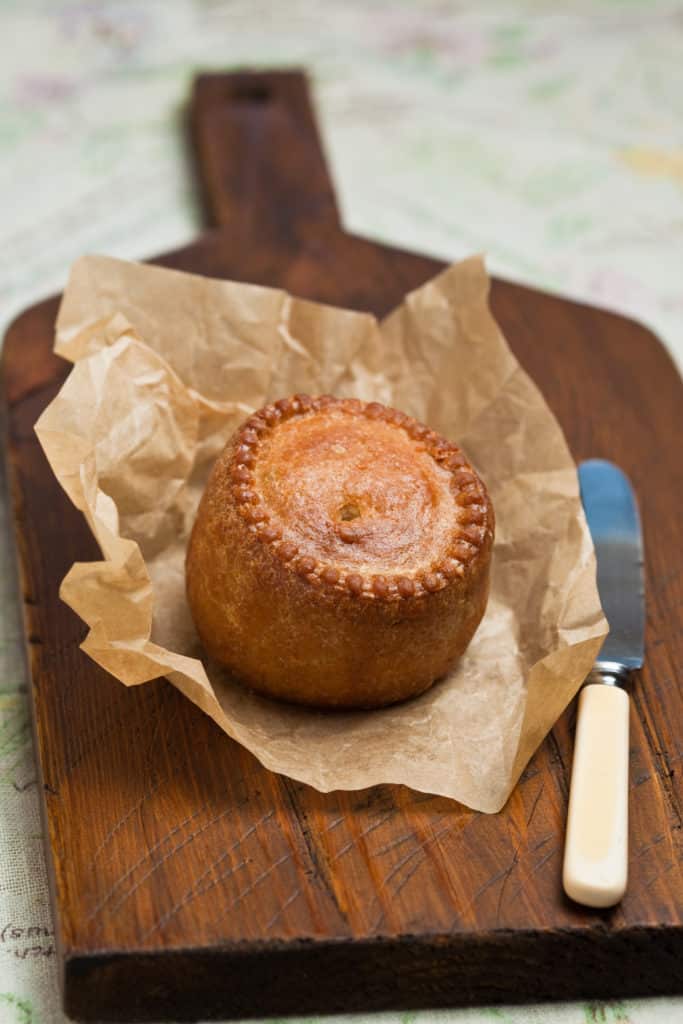 A whole pork pie on a sheet of baking paper which sits on a dark wooden chopping board with a knife at the side. 