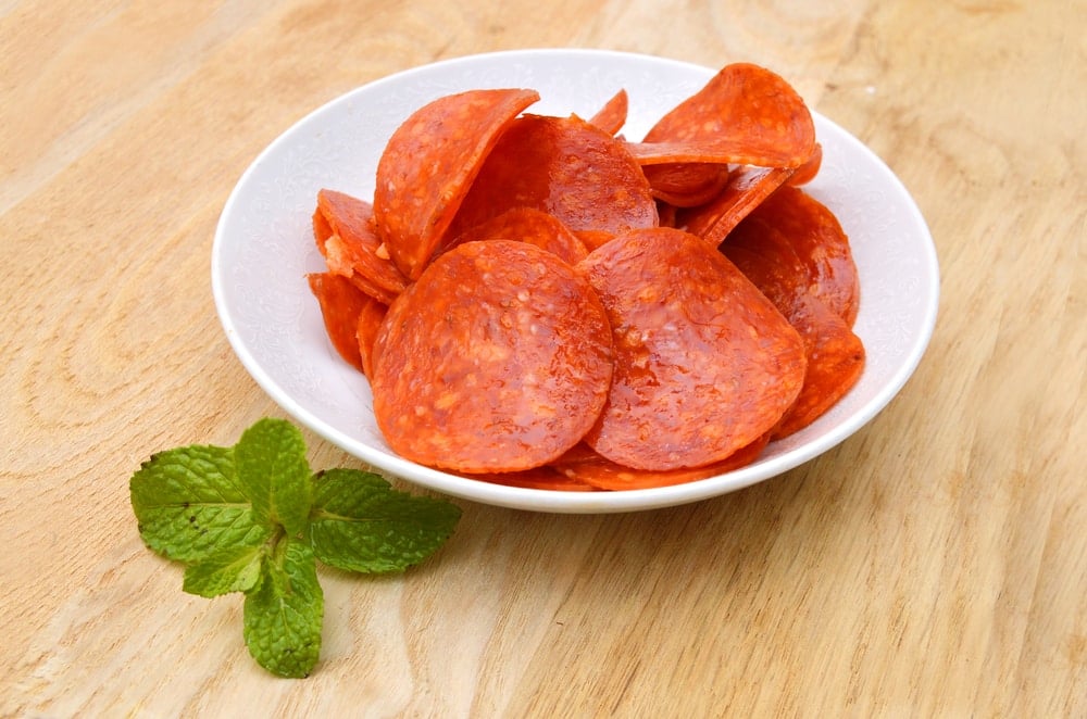 Slices of pepperoni in a white bowl on a wooden table top.