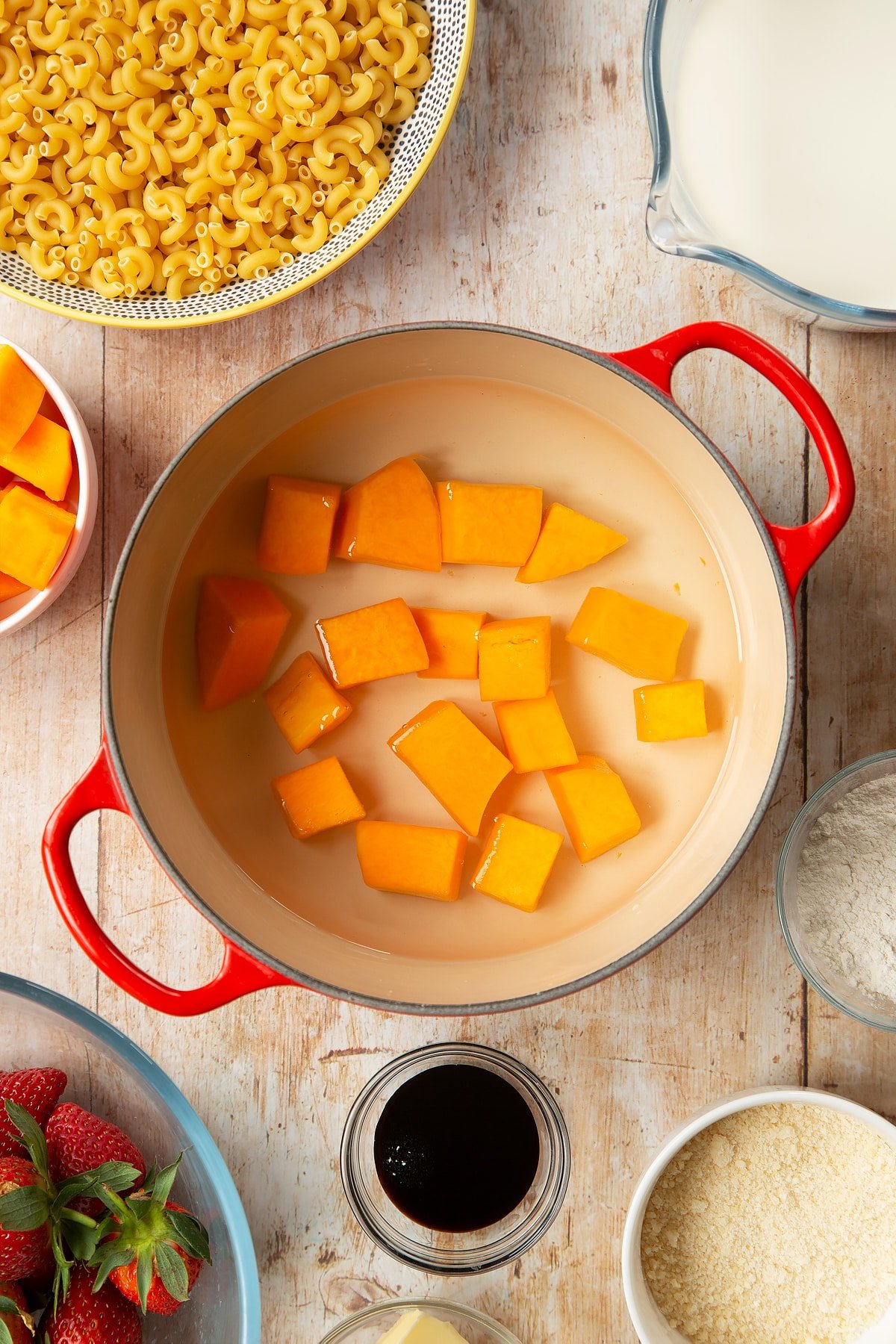 A saucepan containing chunks of butternut squash in water. The pan is surrounded by ingredients for strawberry pasta. 