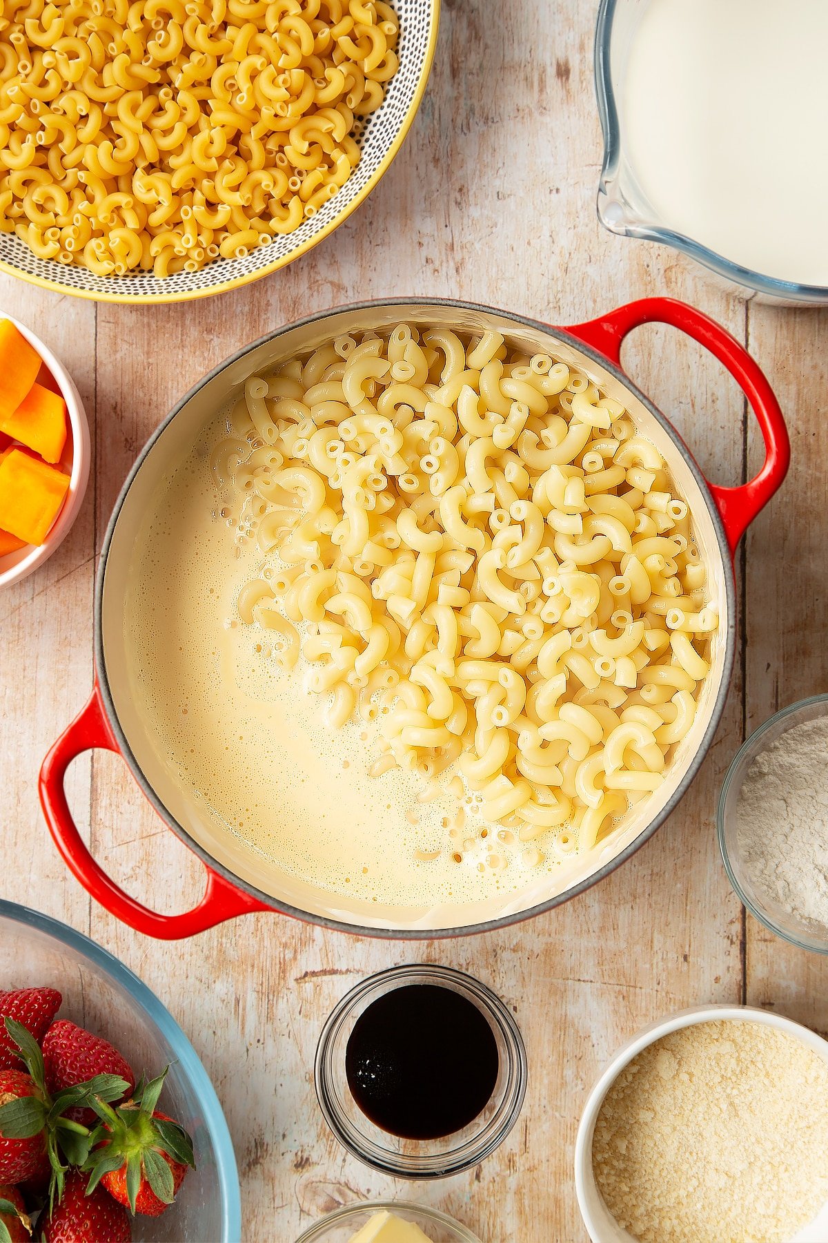 A saucepan containing a cheese and butternut squash sauce, topped with cooked macaroni. The pan is surrounded by ingredients for strawberry pasta. 