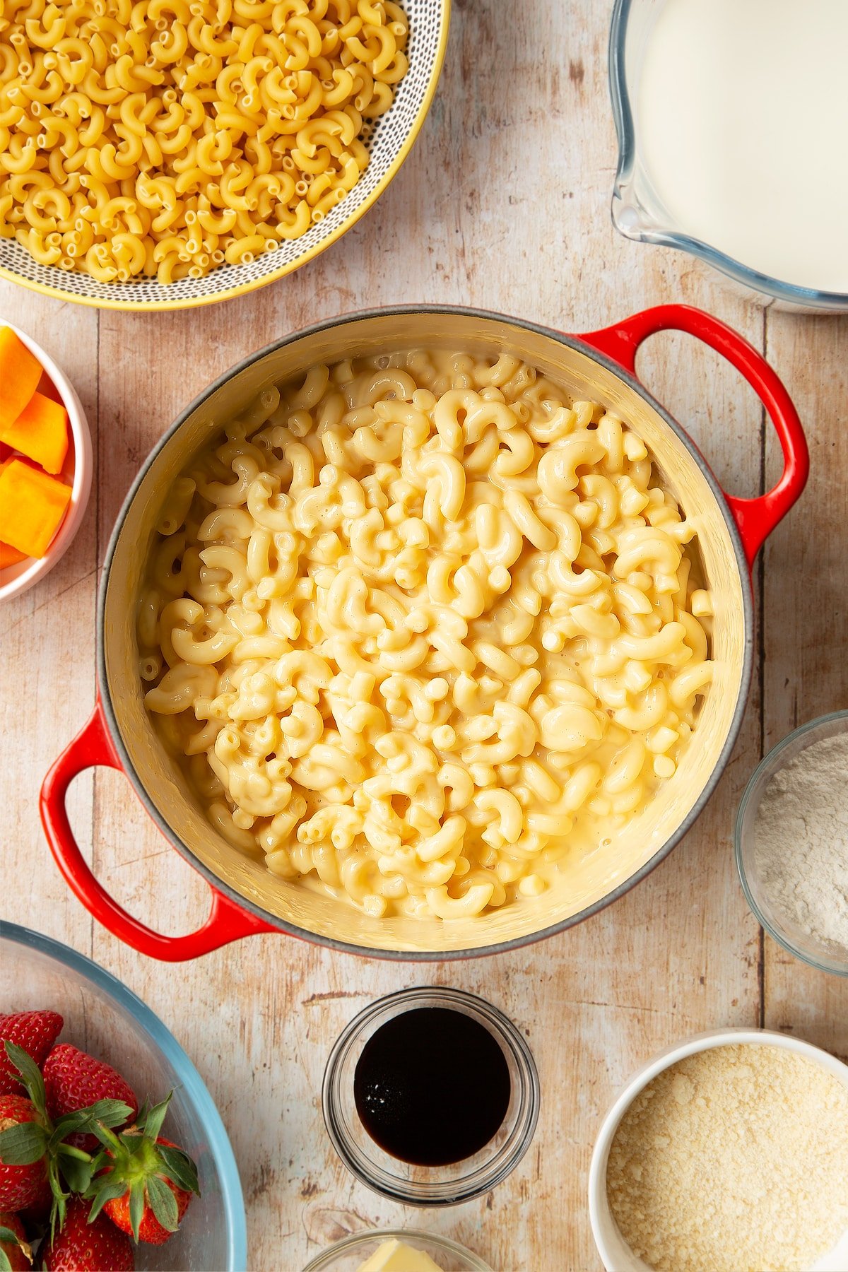 A saucepan containing macaroni stirred into a cheese and butternut squash sauce. The pan is surrounded by ingredients for strawberry pasta. 