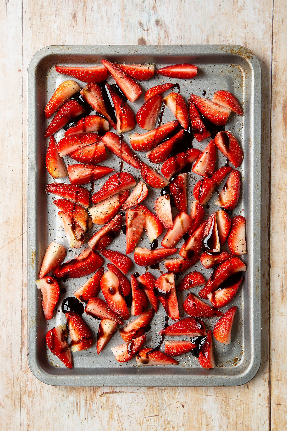 Strawberries sliced into quarters on a non-stick tray, seasoned with pepper and drizzled with balsamic glaze.