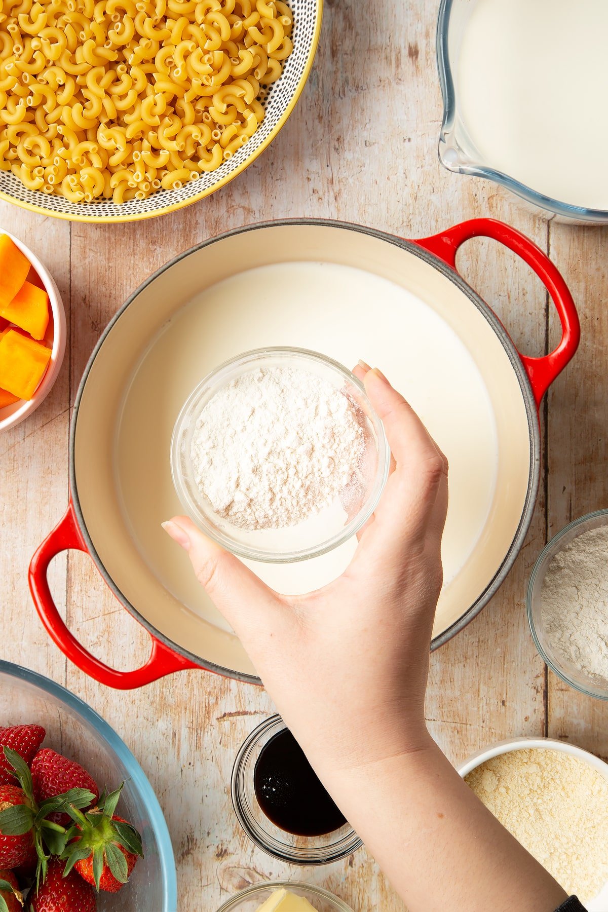 A saucepan containing milk. A hand above it holds a small bowl of flour. The pan is surrounded by ingredients for strawberry pasta. 