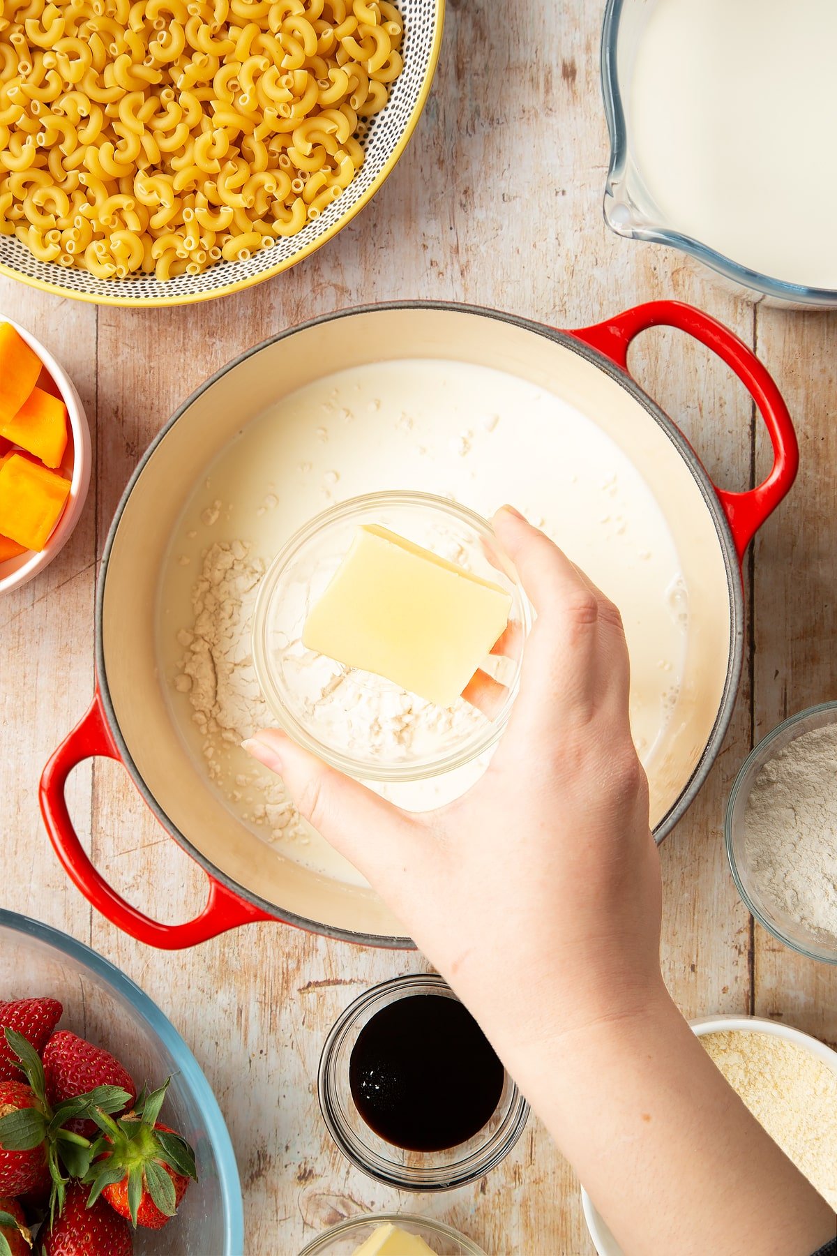 A saucepan containing milk and flour. A hand above it holds a small bowl of butter. The pan is surrounded by ingredients for strawberry pasta. 