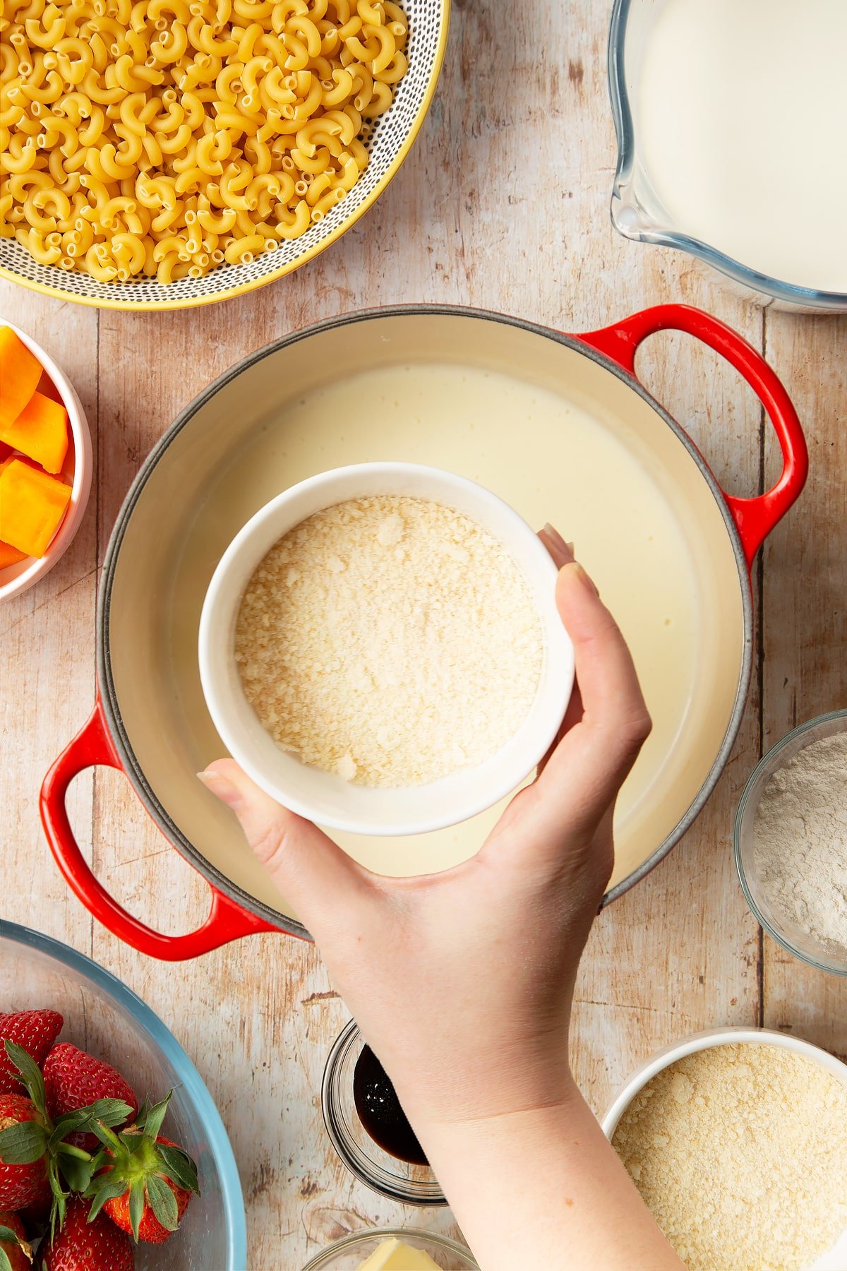 A saucepan containing a white sauce made with milk, butter and flour. A hand above it holds a small bowl of grated Parmesan cheese. The pan is surrounded by ingredients for strawberry pasta. 