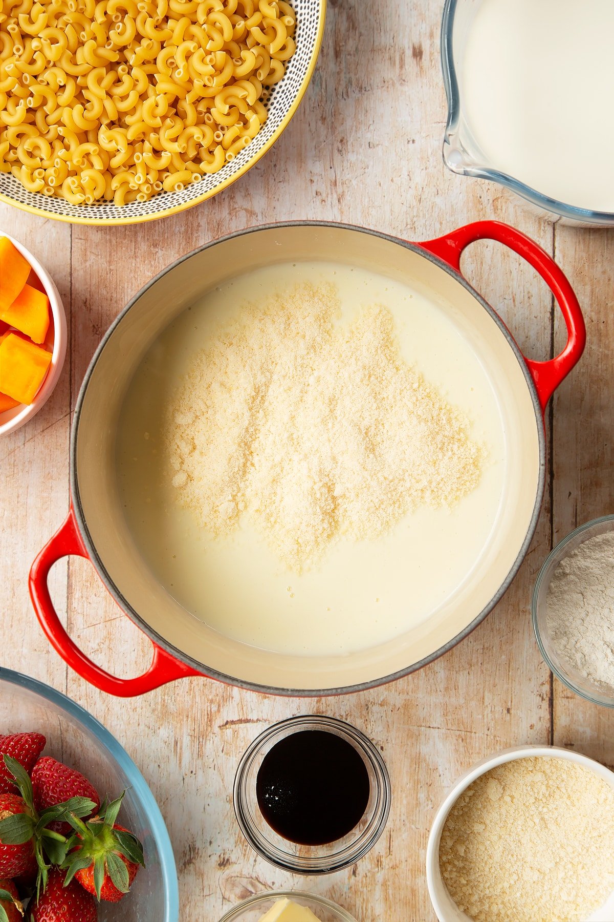 A saucepan containing a white sauce made with milk, butter and flour, topped with Parmesan cheese. The pan is surrounded by ingredients for strawberry pasta. 