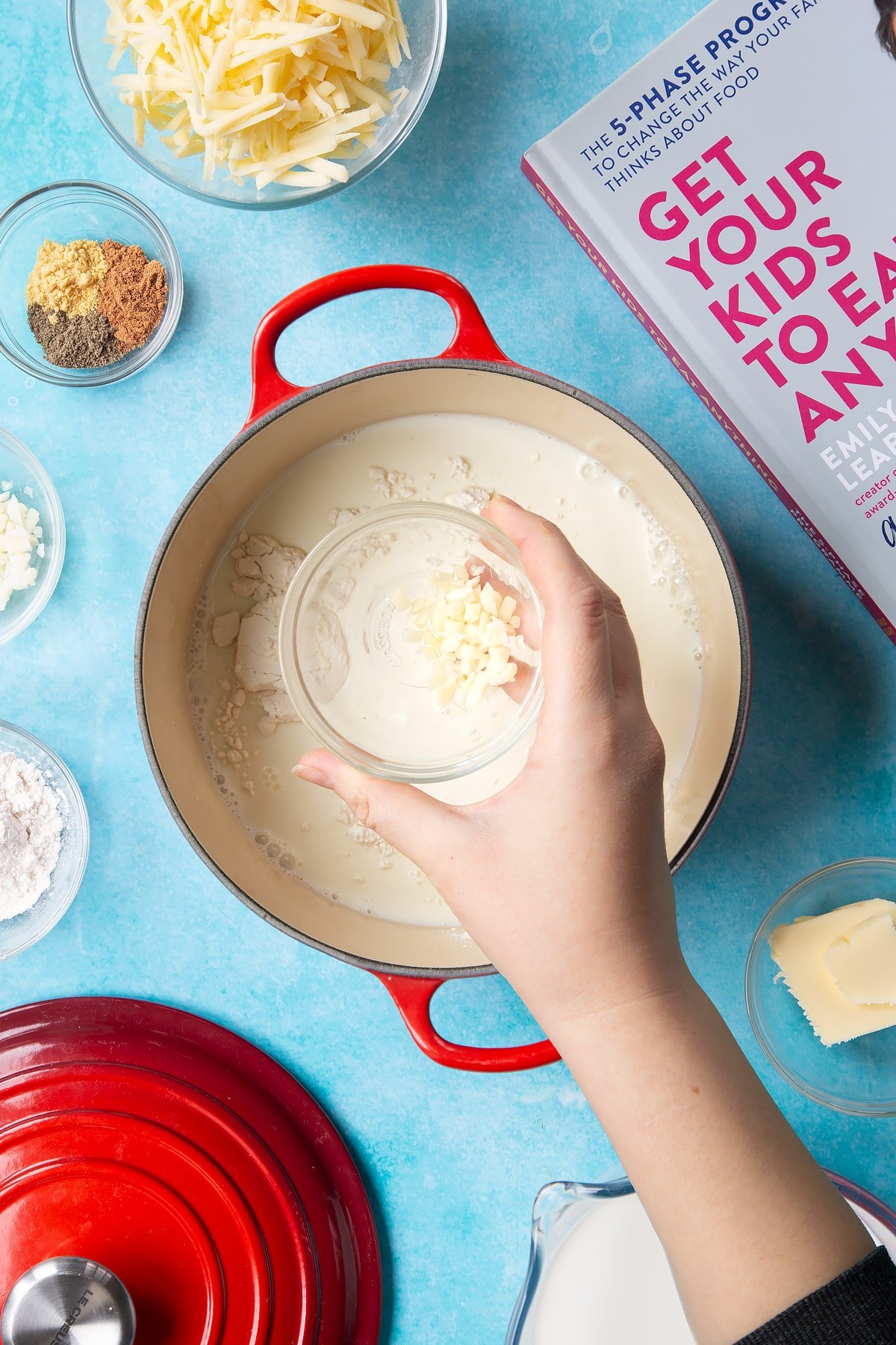 A pan containing milk and flour. A hand holds a small bowl of chopped garlic. The pan is surrounded by ingredients to make cheese sauce for a vegetable fondue.