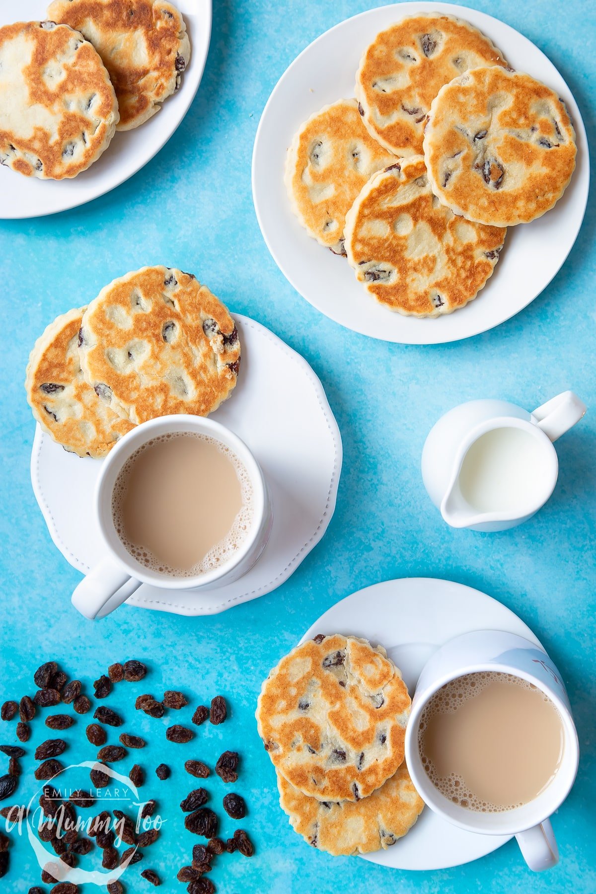 Welsh cakes on a number of white plates with cups of tea. A small jug of milk sits to one side by a scattering of sultanas.