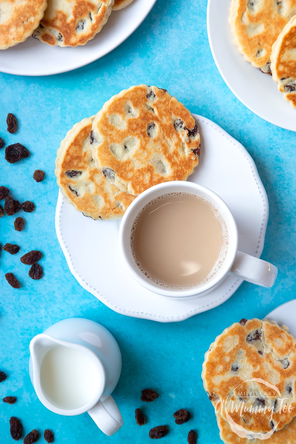 Welsh cakes on a white plate with a cup of tea. A small jug of milk sits to one side by a scattering of sultanas. 