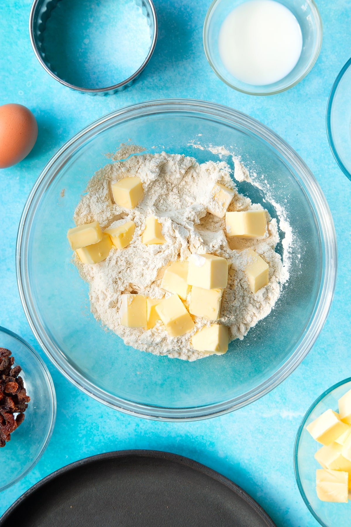 Flour and cubed butter in a bowl. Ingredients and equipment to make Welsh cakes surround the bowl.