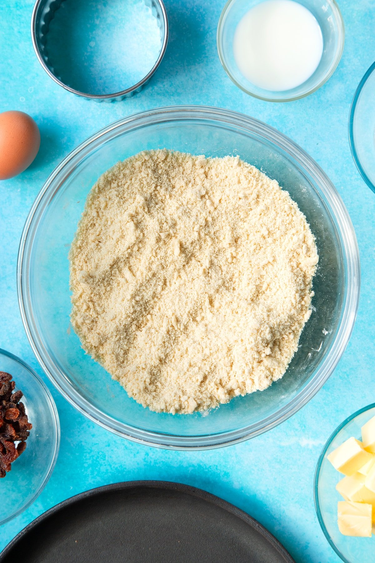 Flour and cubed butter rubbed together to a crumb in a bowl. Ingredients and equipment to make Welsh cakes surround the bowl.