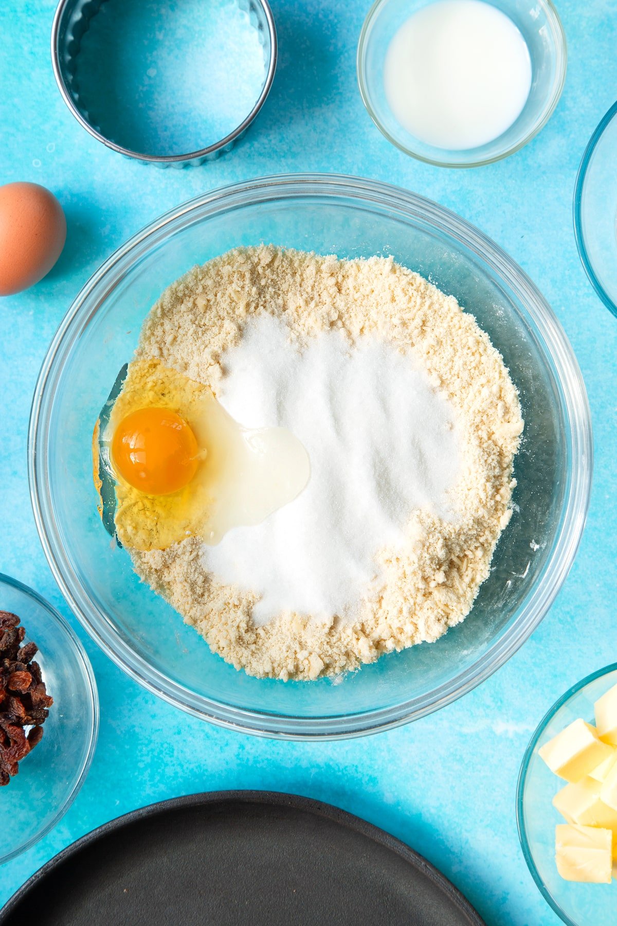 Flour and cubed butter rubbed together to a crumb in a bowl. An egg and sugar sit on top. Ingredients and equipment to make Welsh cakes surround the bowl.