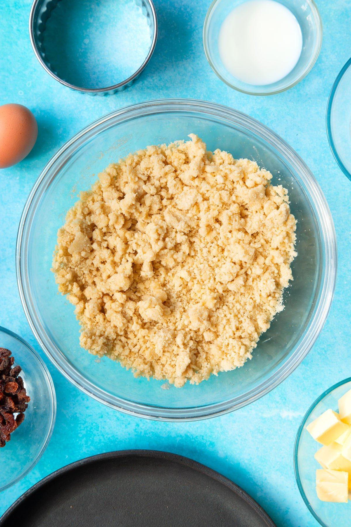 Flour, butter, egg and sugar mixed together in a bowl. Ingredients and equipment to make Welsh cakes surround the bowl.