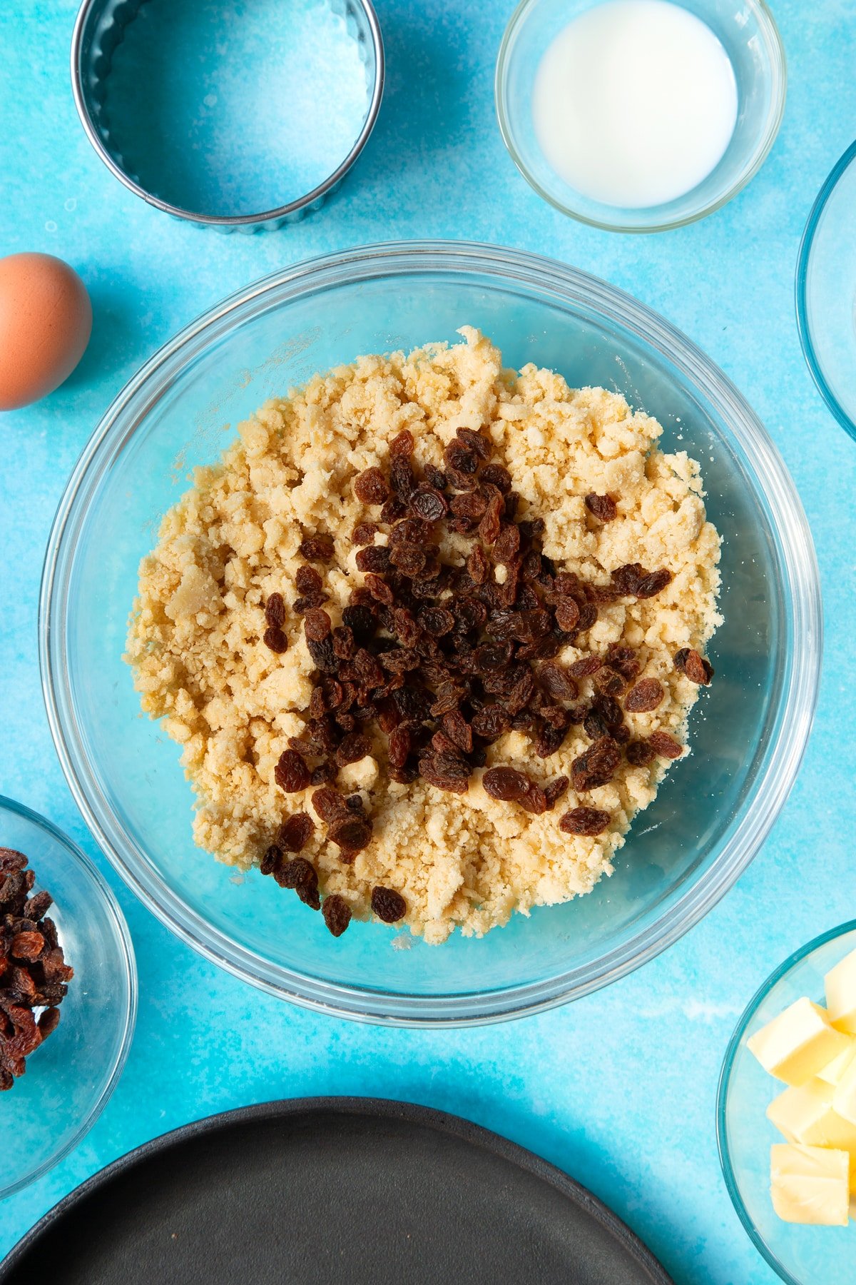 Flour, butter, egg and sugar mixed together in a bowl, with sultanas on top. Ingredients and equipment to make Welsh cakes surround the bowl.