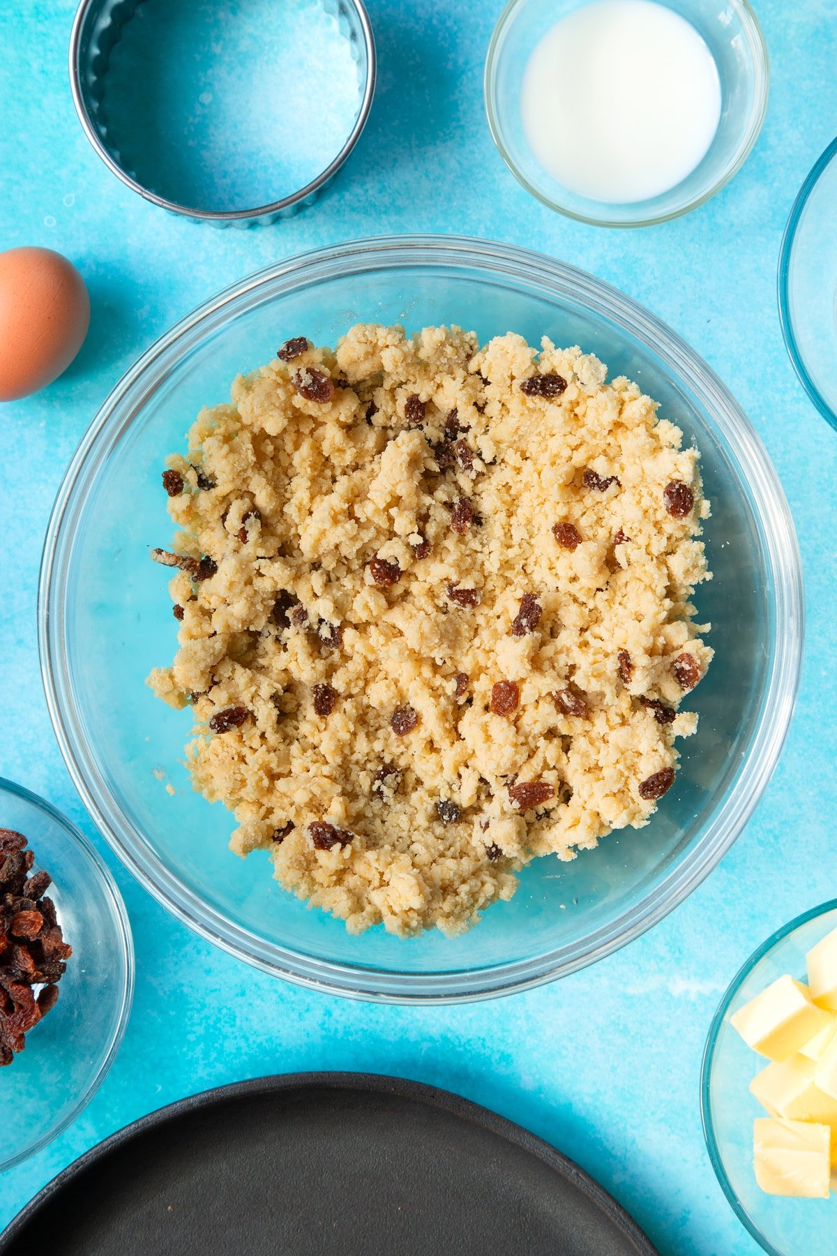 Flour, butter, egg, sugar and sultanas mixed together in a bowl. Ingredients and equipment to make Welsh cakes surround the bowl.