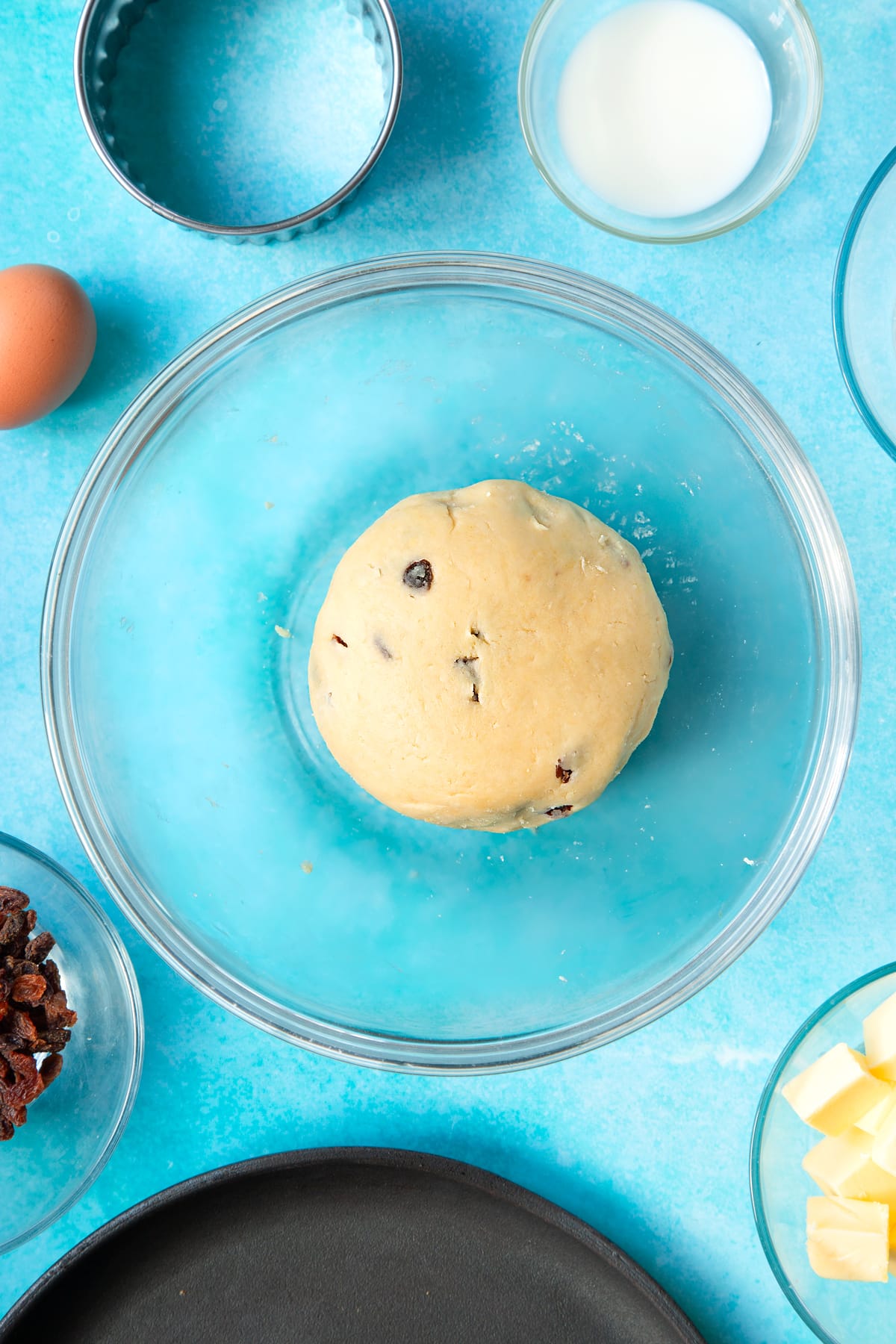 A dough made from flour, butter, egg, sugar and sultanas in a bowl. Ingredients and equipment to make Welsh cakes surround the bowl.