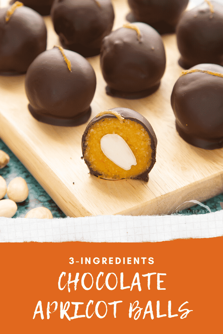 Chocolate apricot balls arranged on a wooden board. In the foreground sits half a ball, revealing the bright orange filling. An almond half sits in the centre of the filling. Caption reads: 3-ingredients chocolate apricot balls.