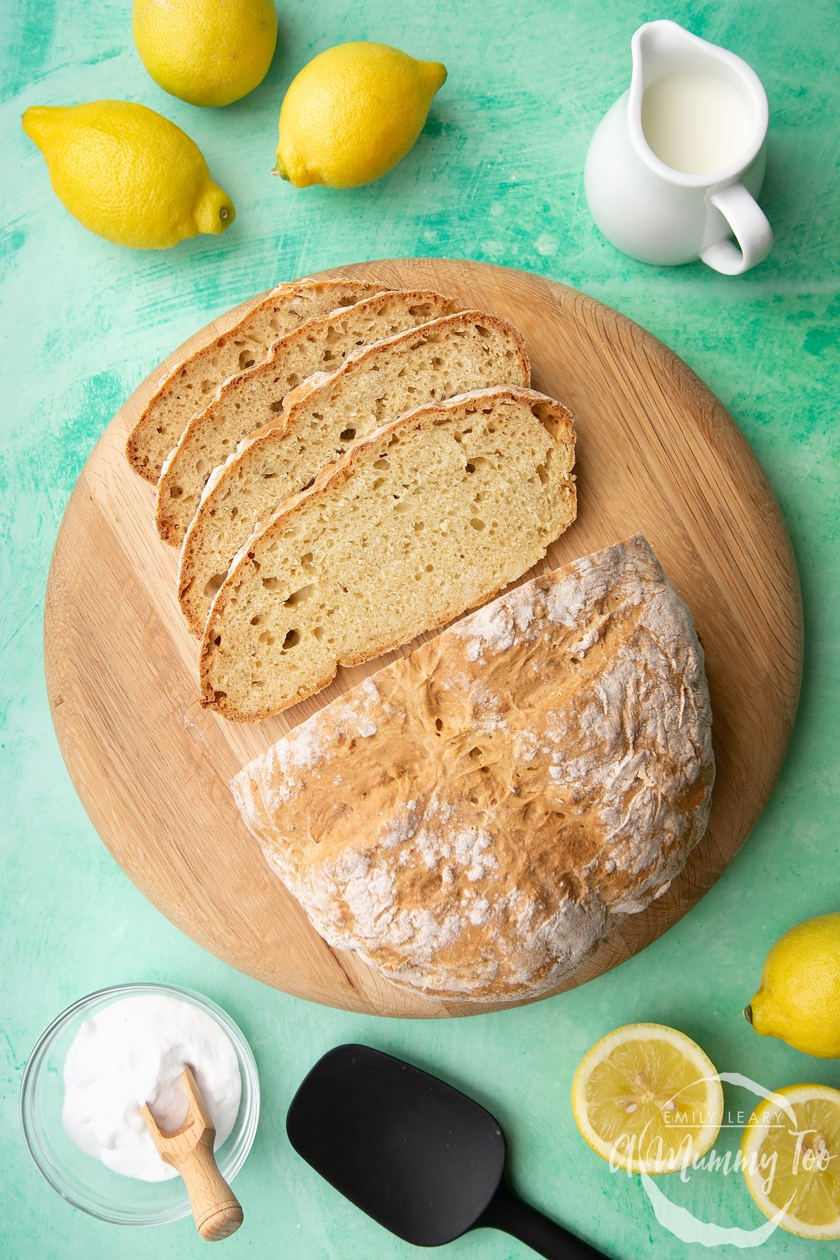 Soda bread without buttermilk on a wooden board. Some of it is sliced. Lemon, milk and bicarbonate of soda are placed around the board.